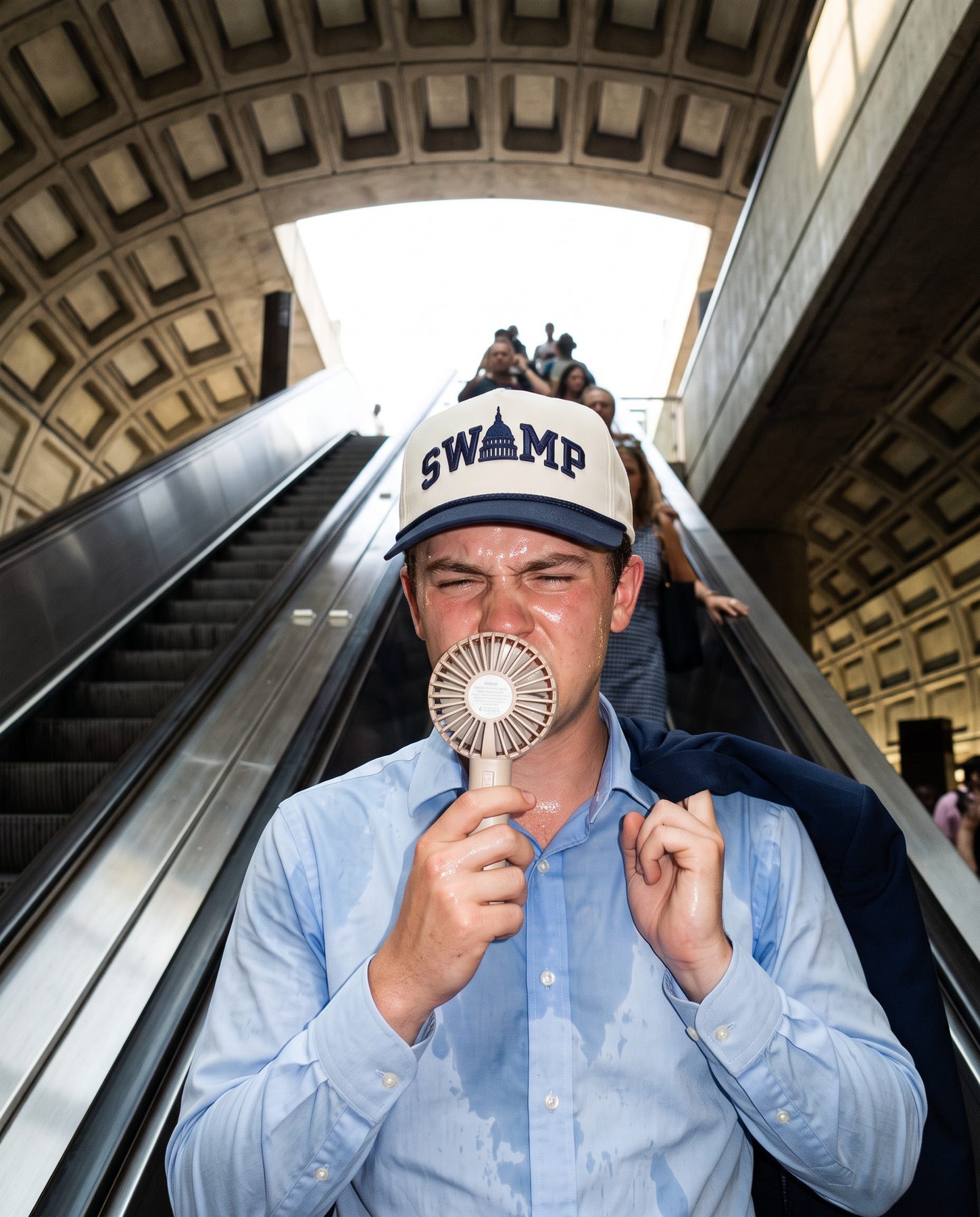 A sweating staffer on a Metro escalator with a tiny fan, wearing The Swamp cap