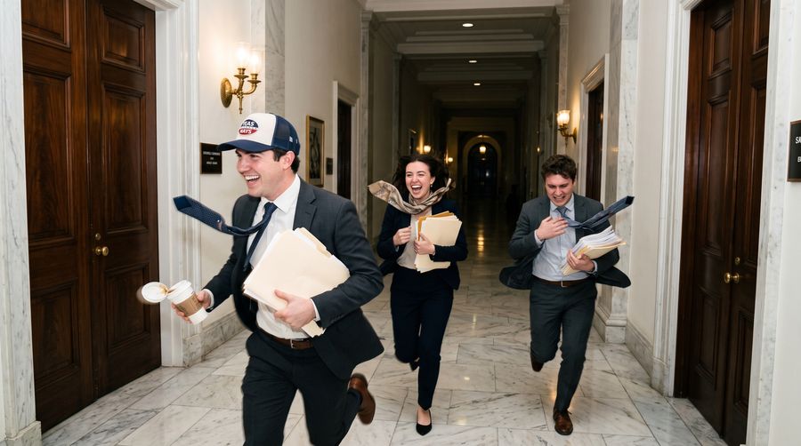 Three staffers sprinting down a marble office building corridor, ties flying, one in the Yeas and Nays trucker