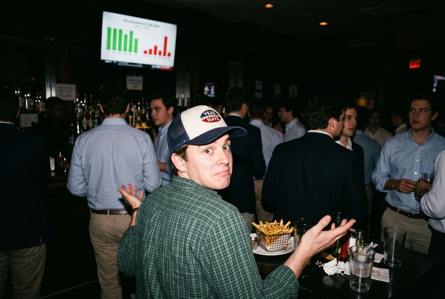 A staffer in the Yeas and Nays trucker shrugging in a crowded Hill bar